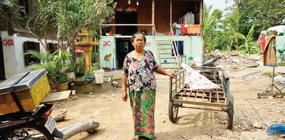 Eine ältere kambodschanische Frau steht vor einem Lastenkarren, sie trägt ein weites Hemd und einen Rock, Sandalen, hinter ihr eine Hütte.