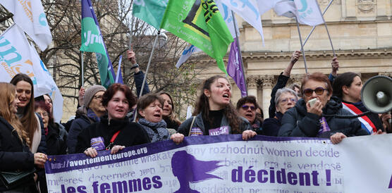 Das Foto zeigt Frauen hinter einem Banner bei einer Demonstration. Eine von ihnen spricht in ein Megafon. Andere schwenken Fahnen.