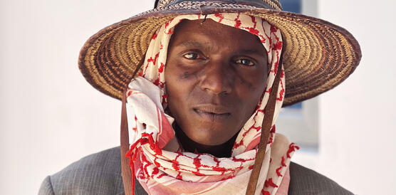 Ein nigerianischer Mann mit Strohhut trägt ein Jacket und ein Kopf-Hals-Tuch.