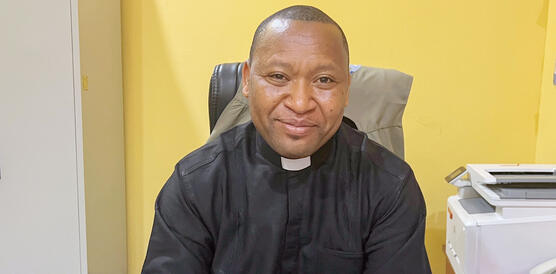 Ein nigerianischer Priester – ein Mann mit kurz rasierten Haaren in Priesterkleidung – sitzt in einem Büro an einem Schreibtisch.