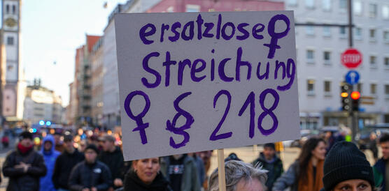 Das Bild zeigt im Hintergrund viele Menschen, im Vordergrund ein Protestschild mit Aufschrift "§218 ersatzlos streichen"