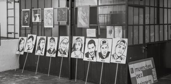 Das Foto zeigt neun Schilder, die in einem Raum auf dem Boden stehen. Die Schilder zeigen jeweils die Porträtsfotos der im Jahr 2016 beim OEZ-Anschlag in München ermordeten Menschen.