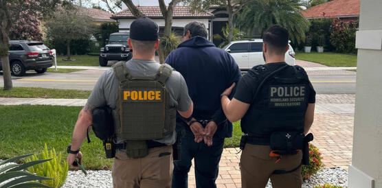 Das Foto zeigt von hinten zwei Männer mit Holster und Schutzwesten, auf denen "Police" steht, wie sie einen Mann abführen, dessen Hände auf dem Rücken mit Handschellen gefesselt sind.