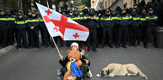 Eine Frau mit georgischer und europäischer Fahne und einem Teddybär sitzt auf der Straße vor einer Reihe Polizisten.