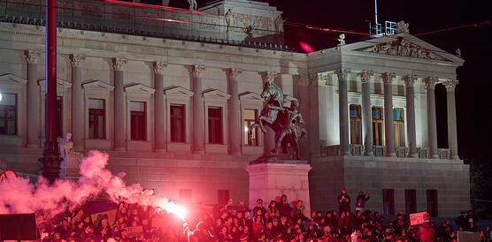 Das Foto zeigt eine demonstrierende Menschenmenge vor einem Gebäude bei Nacht mit Fahnen und Plakaten. Eine Person hat ein Benglo entzündet.
