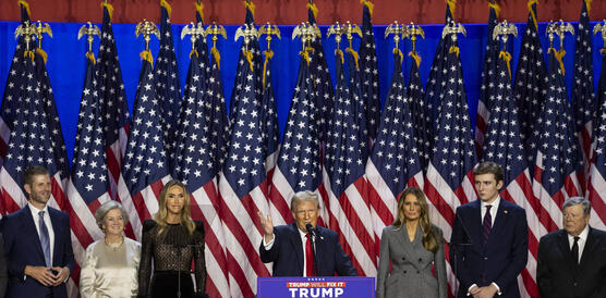 Das Bild zeigt mehrere Menschen auf einer Bühne, ein Mann steht an einem Podium, im Hintergrund viele USA-Flaggen