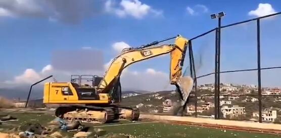 Das Foto zeigt einen Bagger auf einem Fußballplatz. Er reißt mit seiner Schaufel einen hohen Zahn ein.
