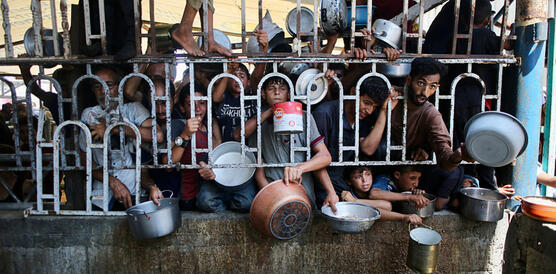 Verzweifelte Menschen drängen sich hinter einem Gitter, durch das einige flehend ihre Töpfe und Teller stecken.