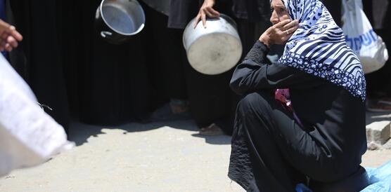 Eine Frau mit Kopftuch sitzt auf dem staubigen Boden neben einer Gruppe Menschen und hat ihr Kinn auf ihre linke Hand gestützt.