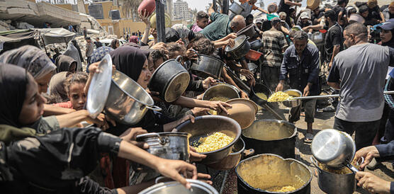 Das Foto zeigt eng zusammenstehende und teilweise übereinander liegende Menschen, die verzweifelt ihre Teller und Töpfe in die Höhe halten und in Richtung der Hilfskräfte, die Essen verteilen.