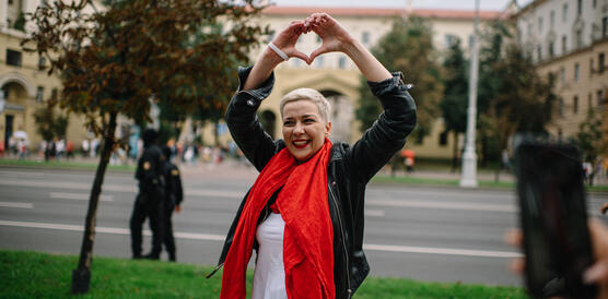 Das Foto zeigt Maryia Kalesnikava. Sie lacht und formt mit ihren beiden Händen, die sue über ihren Kopf hält, ein Herz.