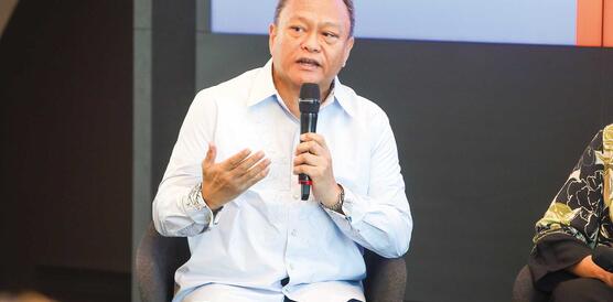 Ein Mann mittleren Alters sitzt in ein Hemd gekleidet auf einer Bühne, hält ein Mikrofon in der linken Hand und gestikuliert mit der anderen.