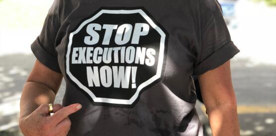 Aufnahme einer Frau an einem sonnigen Tag. Sie blickt leicht lächelnd in die Kamera und zeigt mit ihrer rechten Hand auf ihr T-Shirt, auf dem grop steht: "Stob executions now!"