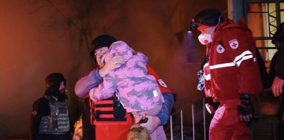 Rettungskräfte tragen ein kleines Kind aus einem Haus. Im Hintergrund lodert Feuer.