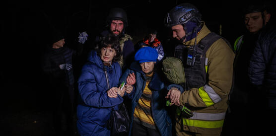 Mehrere Rettungskräfte stehen um eine ältere Frau herum. Einer von ihnen hält ihre Hand. Sie alle sind von Dunkelheit umgeben.