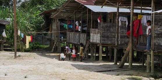 Das Foto zeigt mehrere Holzhütten, errichtet auf niedrigen Stelzen. Auf den Veranden stehen Menschen.