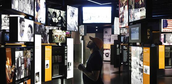 Mann steht in einem Museum vor übereinander- und nebeneinandergehängten Bildschirmen, die verschiedene Szenen schwarzen Lebens in den USA zeigen.