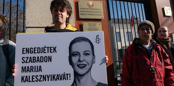 Ein junger Mann steht vor einem Mauerzaun und hält ein Schild in die Kamera, auf dem unter anderem die Amnesty-Kerze und eine Porträtzeichnung von Maryia Kalesnikava abgebildt ist. Neben ihm stehen weitere Personen.