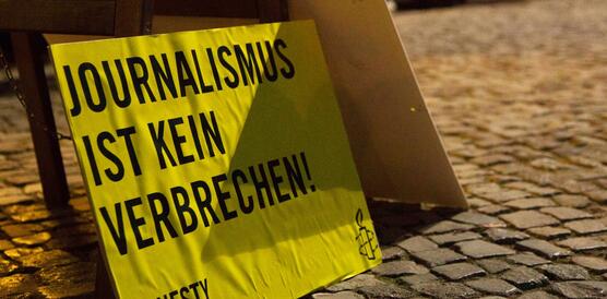 Das Foto zeigt ein Pappschild mit Amnesty-Logo und dem Schriftzug "Journalismus ist kein Verbrechen" auf dem Kopftseinpflaster eines Platzes stehend.