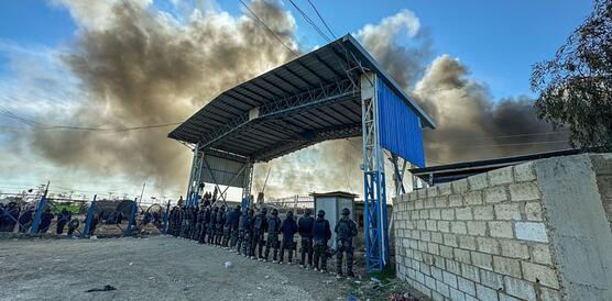Das Foto zeigt mehr als zwanzig uniformierte Sicherheitskräfte mit Helmen, Westen und Schutzschilden von hinten fotografiert. Sie stehen in einer Reihe in einem großen Tor, das in ein Camp führt. Links von ihnen stehen Personen hinter einem Maschendrahtzahn. Rechts von ihnen befindet sich eine kleine Mauer.