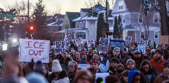 Protestzug mit vielen Menschen zieht durch eine Straße. Die Menschen halten unter anderem Schilder hoch, auf denen steht "ICE out".