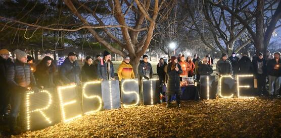 Das Foto zeigt eine größere Gruppe Menschen abends vor einem Baum im Halbkreis stehen. Viele halten Kerzen in der Hand. Vor ihnen steht auf Papptafeln "Resist ICE".
