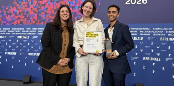 Das Foto zeigt drei Personen, die eng nebeneinander stehen und in die Kamers lächeln. Die Person in der Mitte hält eine Urkunde in der Hand. Auf der Wand hinter ihnen steht unter anderem: 76. Berlinale.