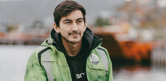 Ein junger Mann mit Schnurrbart trägt Arbeitskleidung und sitzt auf einem Betonblock an einem Hafen.