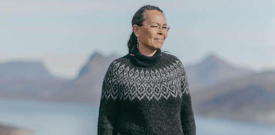 Eine Frau in Turnschuhen, Jeans und Norwegerpullover steht draußen in einer Fjordlandschaft und lächelt.
