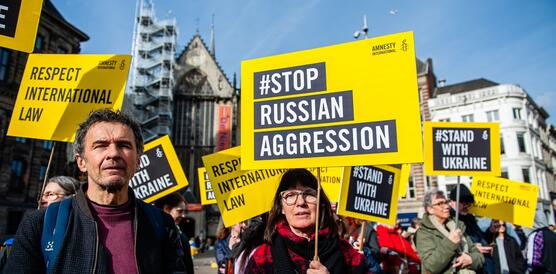 Das Foto zeigt eine Frau neben einem Mann bei einer Demonstration. Die Frau hält ein Schilder mit Amnesty-Logo. Auf dem Schild steht: "Stop Russian Aggression". Im Hintergrund protestieren weitere Personen mit Schildern.