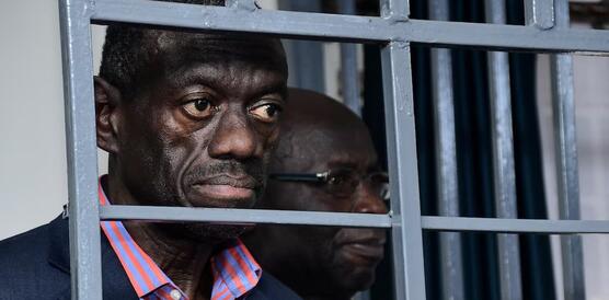 Der ugandische Oppositionspolitiker Dr. Kizza Besigye in einem dunkelblauen Sakko und orange-blau gestreiften Hemd hinter grauen Gefängnisstäben.