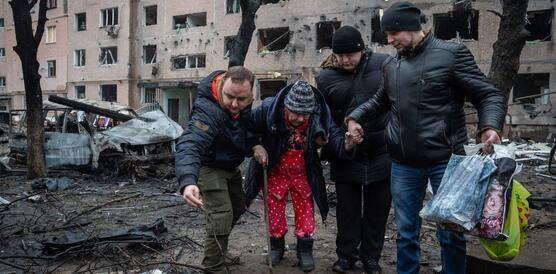 Drei Mäner stützten eine ältere Frau mit Gehstock vor einem zerstörten Gebäude.