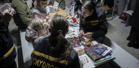 Das Foto zeigt zwei Frauen in Uniform mit zwei Kindern an eine Tisch. Sie spielen. Im Hintergrund stehen weitere Personen.