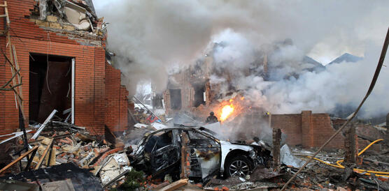 Das Foto zeigt zerstörte Häuser und im Vordergrund ein zerstörtes Auto. Auf der Straße brennt es an einer Stelle. Rauch steigt auf. Überall liegen Trümmer.