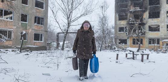 Eine mit zwei Tüten bepackte Frau geht durch den Schnee zwischen zwei zerstörten mehrstöckigen Wohngebäuden.