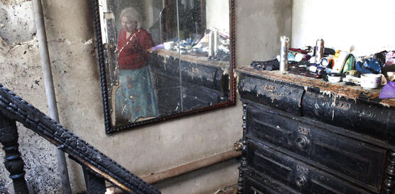 Das Foto zeigt einen Spiegel in einem Hausflur mit angebrannter Treppe und einem angebrannten Schrank. Der Spiegel reflektiert das Bild einer älteren Frau, die im Flur steht.