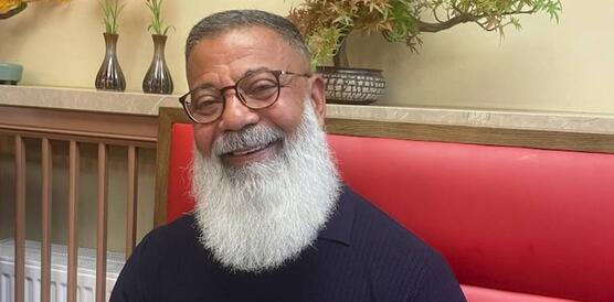 Porträtfoto von Hassan El Sayed, der auf gepolsterten Bank an einem Tisch sitzt. Er hat einen langen Vollbart und trägt Brille und T-Shirt und lächelt in die Kamera.