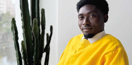 Ein junger Mann aus Ghana trägt Hemd mit Kragen und darüber einen Pullover und sitzt in einem Zimmer, neben ihm eine Kaktuspflanze.