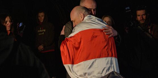 Das Foto zeigt einen in der belarusischen Fahnen gehüllten Mann, der von einem anderen Mann umarmt wird. Um die beiden Männer stehen weitere Personen, von denen einige lächeln.