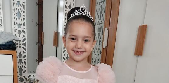 Hind Rajab steht in einem Zimmer. Sie trägt ein Kleid und lächelt in die Kamera. Hinter ihr steht ein Schraank und ein großer Spiegel.k