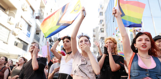 Das Foto zeigt mehrere Personen auf einer Kundgebung. Sie rufen Slogan und halten Regenbogenfahnen in der Hand. Einige lachen.