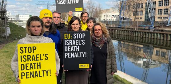 Das Foto zeigt mehrere Personen eng nebeneinander stehen neben einem Kanal. Im Hintergrund steht ein Kran. Sie halten Schilder mit Amnesty-Logos in die Kamera, auf denen steht: "Stop Israel's death Penalty" und "Todesstrafe abschaffen".