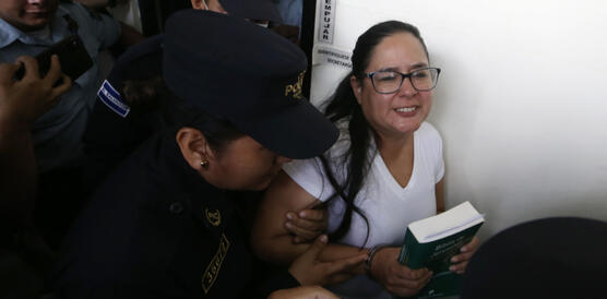 Das Foto zeigt Ruth López, die von uniformierten Personen in Handschellen abgeführt wird. Sie trägt eine Brille und hält ein Buch in ihrer Hand.