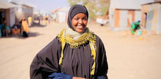 Das Foto zeigt Sadia Moalim Ali auf einer nicht asphaltierten Straße. Sie trägt ein Kopftuch und ein Gewand und lächelt in die Kamera. Im Hintergrund stehen einstöckige Gebäude.