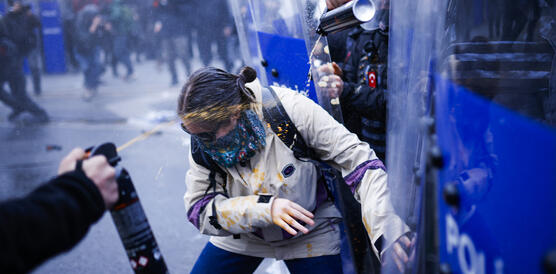 Auf eine vermummte Demonstrantin wird Pfefferspray gespritzt, im Hintergrund Polizei und weitere Demonstrant*innen.
