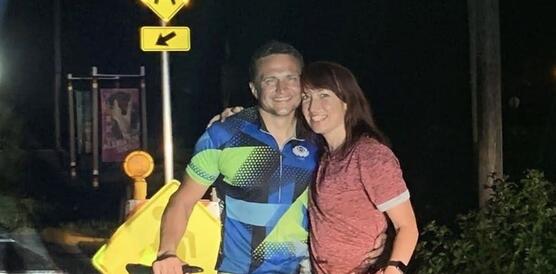 Das Foto zeigt Andriy Shepitsen, der in Sportkleidung hinter einem Fahrrad steht und in die Kamera lächelt. Neben ihm steht eine Frau, die ebenfalls lächelt. Sie umarmen sich gegenseitig. Das Foto wurde abends oder nachts aufgenommen vor einem Verkehrsschild.