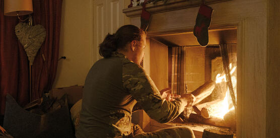 Ein junger Mann mit Bart und langem Haar in Soldatenuniform sitzt vor einem offenen Kamin in dem ein Feuer brennt in einer Wohnung.