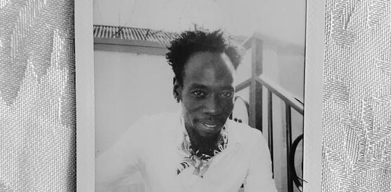 Ein junger Mann aus dem Sudan auf einem Polaroidfoto, das auf einer Tischdecke liegt und auf dem mit Filzstift geschrieben steht: home is a woman