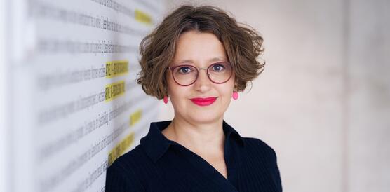 Porträtfoto von Wiebke Gülcibuk, die an eine Wand gelehnt in die Kamera lächelt. Sie trägt eine Brille.