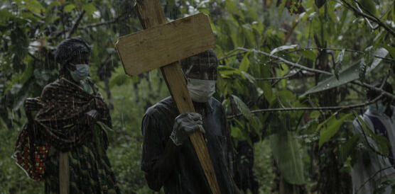 Das Foto zeigt einen Mann mit Mundschutz bei strömenden Regen in einem Dschungel. Er hält ein Holzkreuz in der Hand und schaut in die Kamera.